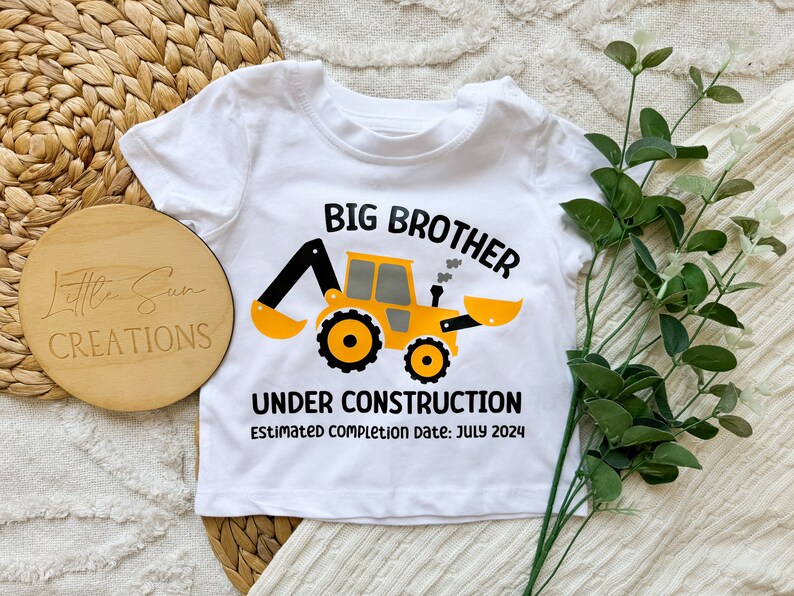 May include: White baby onesie with a yellow construction vehicle graphic and the text "Big Brother Under Construction Estimated completion date: July 2024".