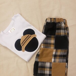 May include: White short-sleeved shirt with a black and brown Mickey Mouse applique. The shirt is paired with brown and black plaid shorts.