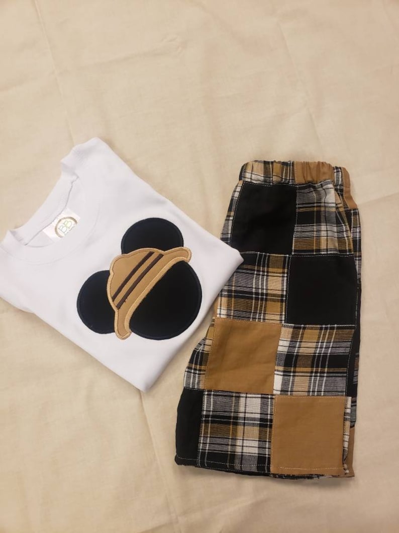 May include: White short-sleeved shirt with a black and brown Mickey Mouse applique. The shirt is paired with brown and black plaid shorts.