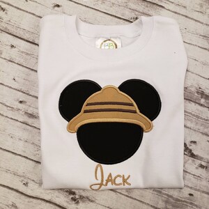 May include: White t-shirt with an embroidered image of Mickey Mouse wearing a safari hat. The name "Jack" is embroidered below the image.