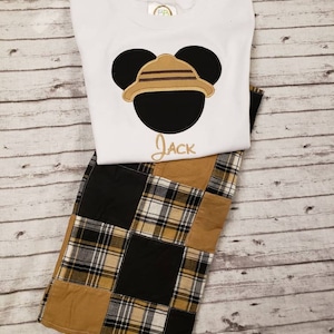 May include: White t-shirt with a black and brown embroidered Mickey Mouse wearing a safari hat. The shirt has the name "Jack" embroidered below the image. The shirt is paired with brown, black, and white plaid shorts.