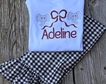 May include: A white long-sleeve shirt featuring an embroidered design of two gray elephants and a dark red bow above the name Adeline in dark red script. The shirt is paired with black and white gingham print pants with ruffled hems.