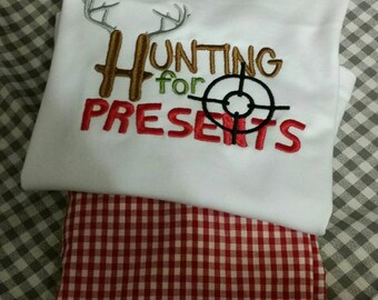 Op de afbeelding: Een wit longsleeve shirt met rode en bruine geborduurde tekst die "Hunting for Presents" zegt met een hertengewei-ontwerp en een doelwitsymbool. Het shirt wordt gecombineerd met een rood-wit geblokte broek.
