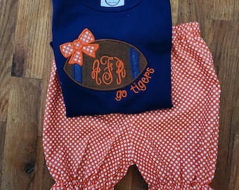 May include: A navy blue short-sleeve shirt featuring an embroidered brown football with a white polka dot bow, a monogram, and the text "go tigers" in orange, paired with orange and white polka dot bloomers with ruffled leg openings.