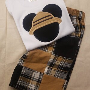 May include: A white t-shirt with a black and brown Mickey Mouse head design. The Mickey Mouse head has a brown hat with stripes. The shirt is paired with brown and black plaid shorts.