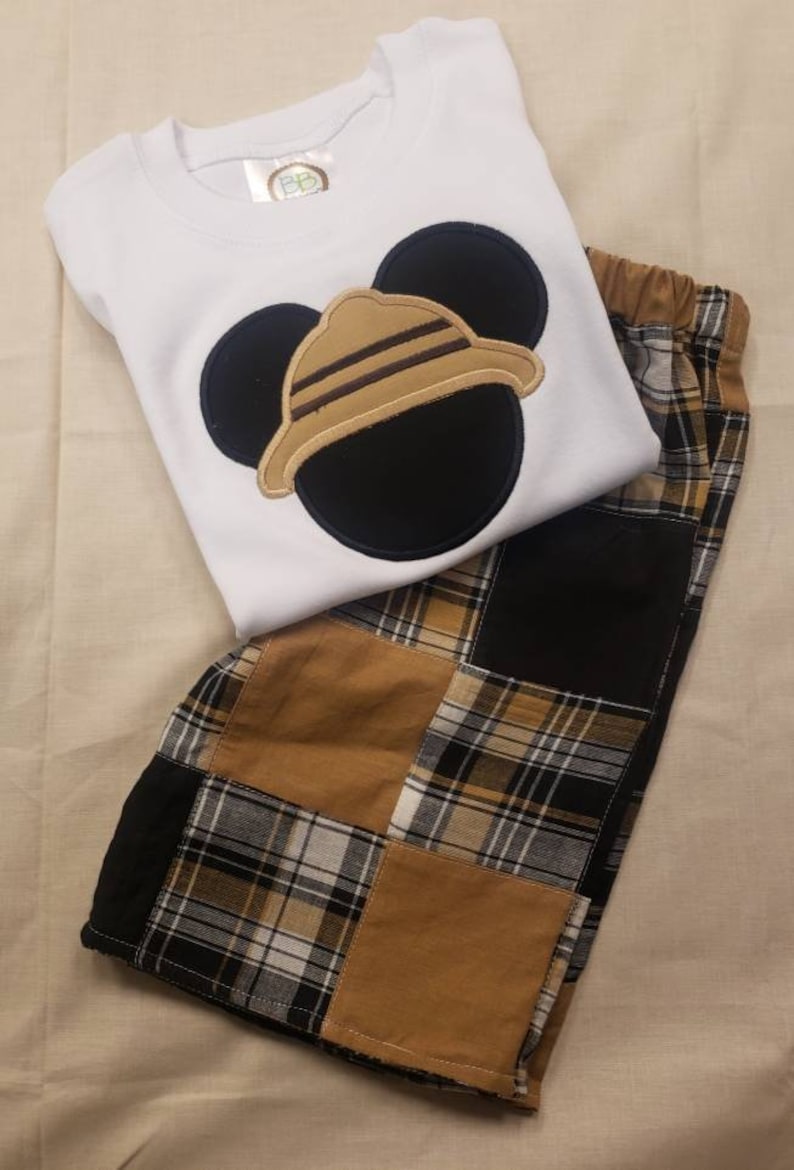 May include: A white t-shirt with a black and brown Mickey Mouse head design. The Mickey Mouse head has a brown hat with stripes. The shirt is paired with brown and black plaid shorts.