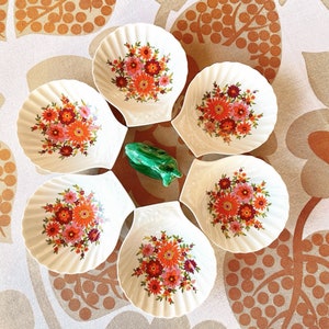 Pode incluir: Seis tigelas de cerâmica branca em forma de concha do mar com um design floral em tons de laranja, vermelho e rosa. As tigelas estão dispostas em círculo sobre um fundo de tecido estampado.
