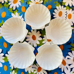 Peut inclure: Cinq bols en céramique blanche en forme de coquillages. Les bols sont disposés en cercle sur un fond à motifs floraux.