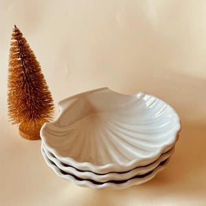 Op de afbeelding: Een stapel van drie witte, schelpvormige schaaltjes met een golvende rand. Links van de schaaltjes staat een kleine, goudkleurige kerstboom. De schaaltjes hebben een glanzende afwerking.