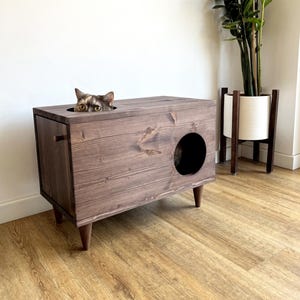 May include: A dark brown wooden cat litter box with a hidden compartment and a round entrance hole. The litter box is on four legs and has a cat peeking out of the top opening.