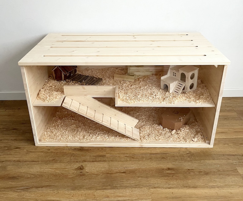 May include: A wooden hamster cage with two levels and a ramp. The cage has a clear plastic front and is filled with wood shavings. There are two wooden houses and a wooden ladder inside the cage.