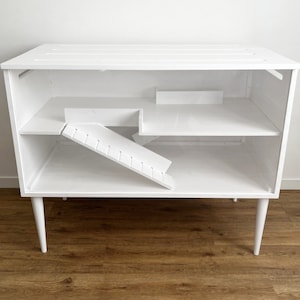 May include: White wooden cage with a ramp and shelves, designed for small animals. The cage has a modern design with white legs.