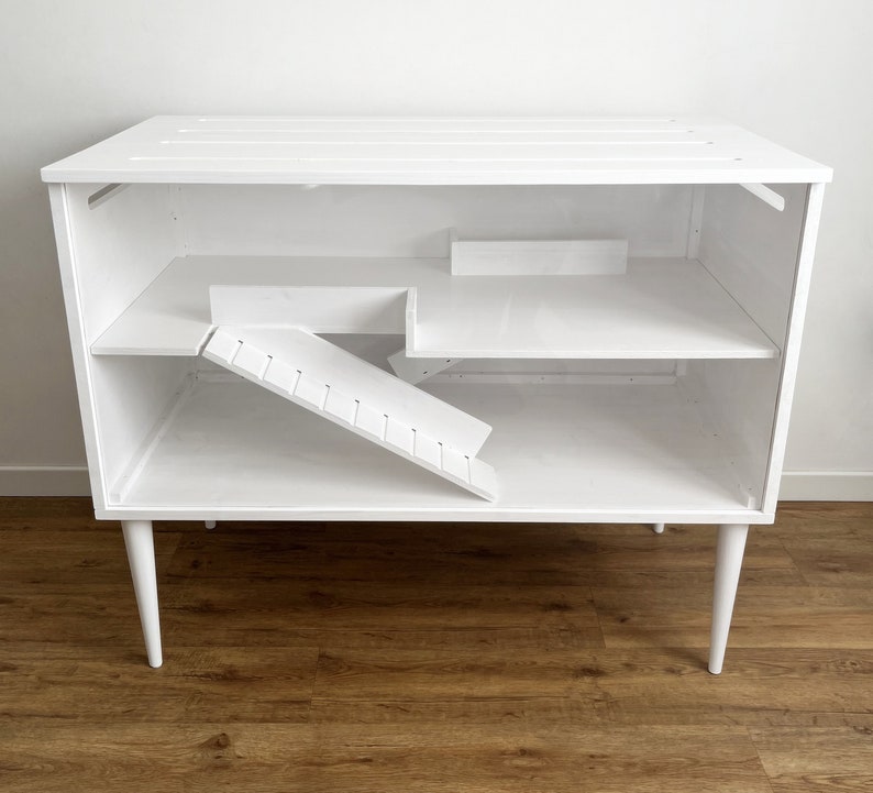 May include: White wooden cage with a ramp and shelves, designed for small animals. The cage has a modern design with white legs.