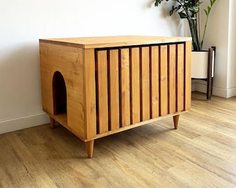 May include: A wooden cat house with a natural finish. The rectangular structure features a rounded entrance on one side and a slatted door on the front. The cat house is elevated on short, angled legs and is designed to blend with home decor.