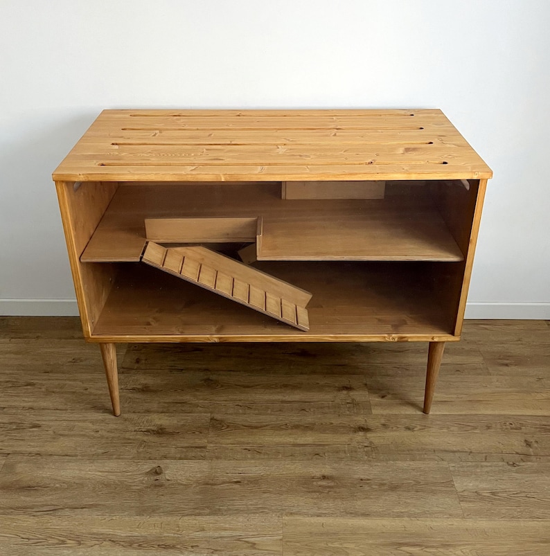 May include: A wooden pet cage with a ramp and a shelf. The cage is made of natural wood and has a light brown finish. The cage is on a wooden floor.