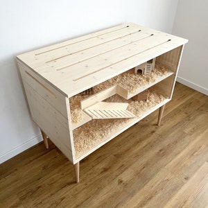 May include: A wooden rabbit hutch with two levels and a ramp. The hutch is made of natural wood and has a light brown finish. The hutch is filled with wood shavings and has a small wooden house inside.