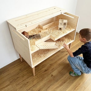 May include: A wooden hamster cage with two levels and a ramp. The cage has a clear plastic lid and is filled with wood shavings. A small wooden house is inside the cage. A young boy is looking at the cage.