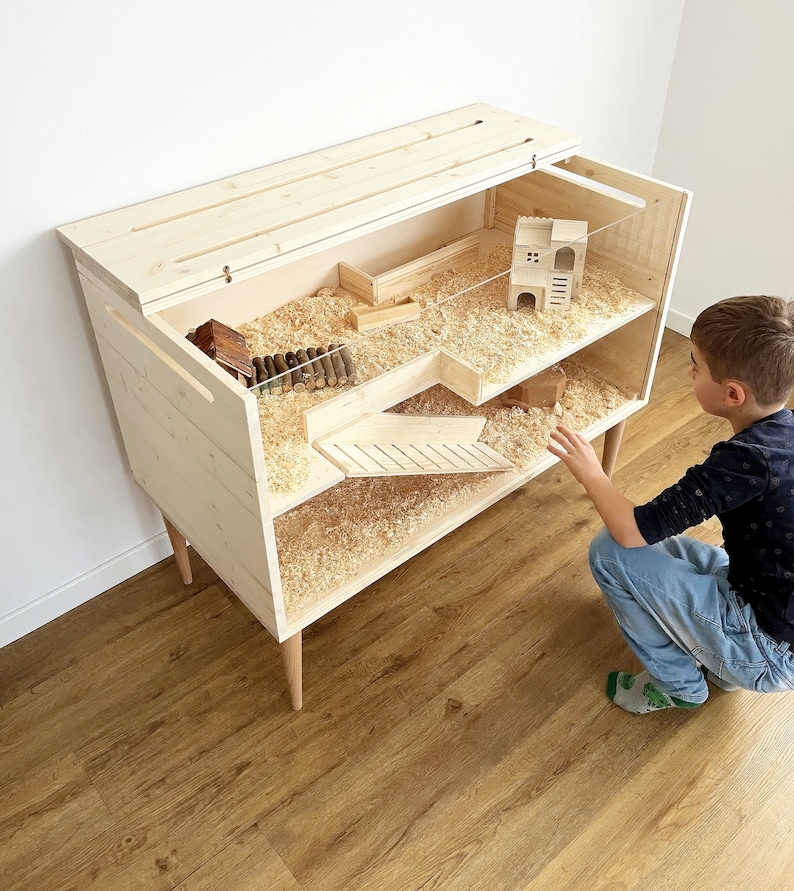 May include: A wooden hamster cage with two levels and a ramp. The cage has a clear plastic lid and is filled with wood shavings. A small wooden house is inside the cage. A young boy is looking at the cage.
