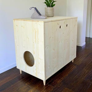 May include: A light-colored wooden cabinet with a rectangular frame, four tapered legs, and a large circular opening on the side. The front features two doors with small circular cutouts for handles. A small plant and a phone stand sit on top.