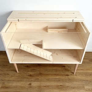 May include: A wooden pet cage with a ramp and a shelf. The cage is made of natural wood and has a clear plastic front. The cage is on four legs.