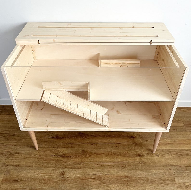 May include: A wooden pet cage with a ramp and a shelf. The cage is made of natural wood and has a clear plastic front. The cage is on four legs.