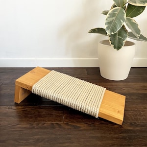 May include: A rectangular wooden ramp with a natural light brown finish, featuring a thick, beige rope tightly wrapped around the center section. The ramp is angled upward, supported by a short wooden block at one end, creating a sturdy incline.