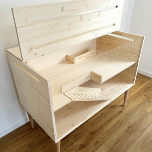 May include: A wooden pet cage with a hinged top and a ramp. The cage is made of natural wood and has a light brown finish. The cage is on four legs and has a clear plastic front.