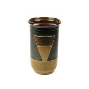 Puede incluir: Un vaso cilíndrico de cerámica que presenta una sección superior esmaltada en gris oscuro y negro con un borde marrón liso. La parte inferior tiene un acabado en esmalte beige mate, acentuado por un diseño central de triángulo invertido en marrón.