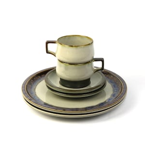May include: Two stacked ceramic coffee cups with brown and white glaze, sitting on a saucer with a brown and blue rim.