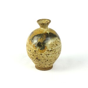 May include: A small, round ceramic vase with a narrow neck and flared rim. The surface features a speckled beige and tan base with a prominent dark brown and black glaze pattern across the upper section, creating a textured, earthy appearance.