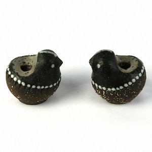 Könnte beinhalten: Zwei kleine schwarze Keramik-Vogelfiguren mit weißen Akzenten. Die Vögel sind in entgegengesetzte Richtungen gerichtet und haben ein Loch oben auf dem Kopf.