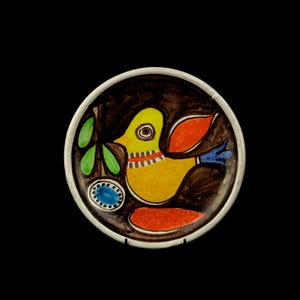 May include: A round ceramic plate featuring a dark brown background with a central yellow bird illustration. The bird has an orange wing, a blue tail, and a red and white striped neck. Green leaves, a blue oval, and an orange shape are also present.