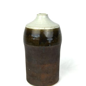 May include: A ceramic jug with a dark brown base and a lighter brown upper section, topped with a cream-coloured rim. The jug has a classic, rustic design, suitable for home decor or storage.