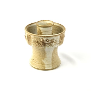 May include: A white ceramic candle holder with a textured design. The holder has a round base and a cylindrical top with a small opening for a candle.