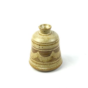 May include: A small, bell-shaped ceramic vase with a speckled cream and tan glaze. The body features a series of horizontal, wavy, incised lines encircling the lower portion, creating a textured pattern against the smooth, mottled surface finish.