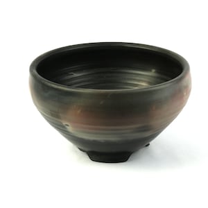 May include: A handcrafted ceramic bowl with a dark, textured exterior. The bowl features a rounded shape with a wide opening and a slightly raised base. The color transitions from dark brown to reddish-brown.