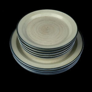 May include: A stack of six beige ceramic plates with a dark blue rim. The plates are stacked in a circular pattern.