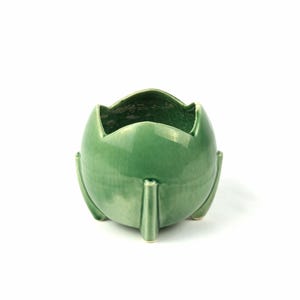 May include: A sage green ceramic planter with a glossy finish, featuring a rounded body and three distinct vertical legs that extend from the base to the rim. The top edge has a scalloped design with four pointed petal-like protrusions.