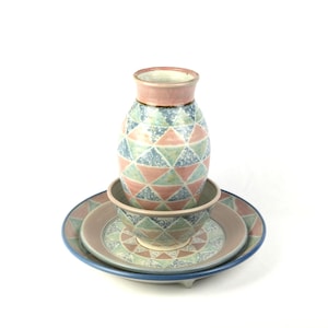 Könnte beinhalten: Ein Keramik-Vasen-, Schalen- und Plattenset. Die Vase hat einen rosa Rand und ein geometrisches Muster in Blau, Grün und Pfirsich. Schüssel und Teller haben passende Muster und Farben. Die Teller sind gestapelt, mit einem blauen Rand auf dem unteren Teller.