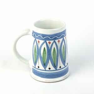 Puede incluir: Una taza de cerámica blanca con un diseño floral azul y verde. La taza tiene un asa y una base redondeada.