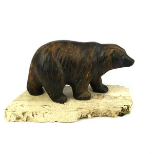 Könnte beinhalten: Eine handgeschnitzte Holzfigur eines Bären mit einer dunkelbraunen und schwarzen Oberfläche. Der Bär steht auf einem hellbraunen Holzsockel.