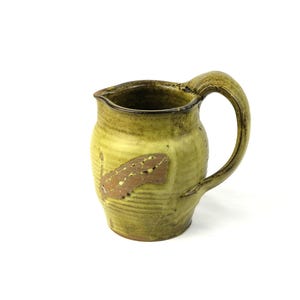 Puede incluir: Jarra de cerámica hecha a mano con esmalte verde y marrón. Tiene un asa curva y un pico vertedor. Un diseño abstracto marrón y beige adorna la parte delantera. Adecuada para servir bebidas. Mide aproximadamente 15 cm de altura.