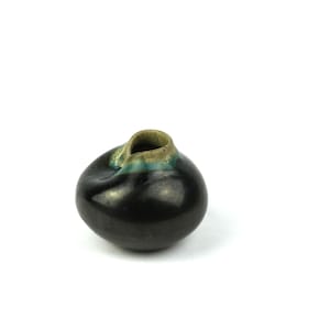 Könnte beinhalten: Kleine, dunkle Keramikvase mit rundem Körper und einzigartiger, gefalteter Öffnung. Die Vase hat eine glänzende schwarze Oberfläche mit einem Hauch von Grün und Gold am Rand. Minimalistisches Design vor weißem Hintergrund.