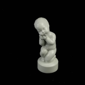 May include: A white porcelain figurine of a baby kneeling with its hands clasped together, looking down. The figurine is on a white pedestal.