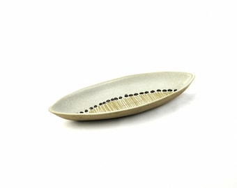 May include: A ceramic, oval-shaped serving dish. The dish has a light beige exterior and a white interior. A line of black dots and a series of parallel lines in beige decorate the interior. The dish is ideal for serving appetizers or small dishes.