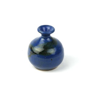 Puede incluir: Un pequeño jarrón redondo de cerámica azul con cuello estrecho y diseño moteado verde oscuro y negro. El jarrón mide aproximadamente 10 cm de alto y tiene una forma sencilla y elegante, adecuado para exhibir flores pequeñas o como elemento decorativo.