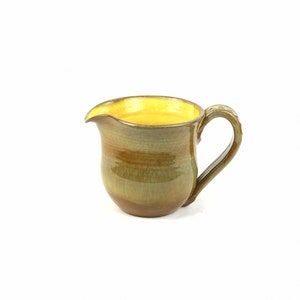 Puede incluir: Un pequeño creamer de cerámica amarillo y verde con asa. El creamer está esmaltado en un color amarillo y verde moteado.
