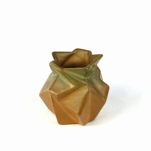 May include: A small, geometric ceramic vase with a gold and green glaze. The vase has a unique, folded design.