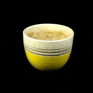 Puede incluir: Un pequeño macetero de cerámica blanca con un esmalte amarillo en la mitad inferior. El macetero tiene una superficie texturizada y una forma ligeramente redondeada.