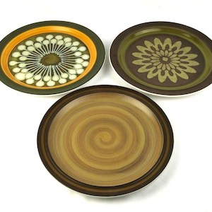 Puede incluir: Tres platos de cerámica con diseños decorativos. Uno presenta un patrón en espiral en tonos marrones y beige. Los otros dos tienen patrones florales y abstractos en verde, naranja y marrón. Los platos están sobre un fondo blanco.